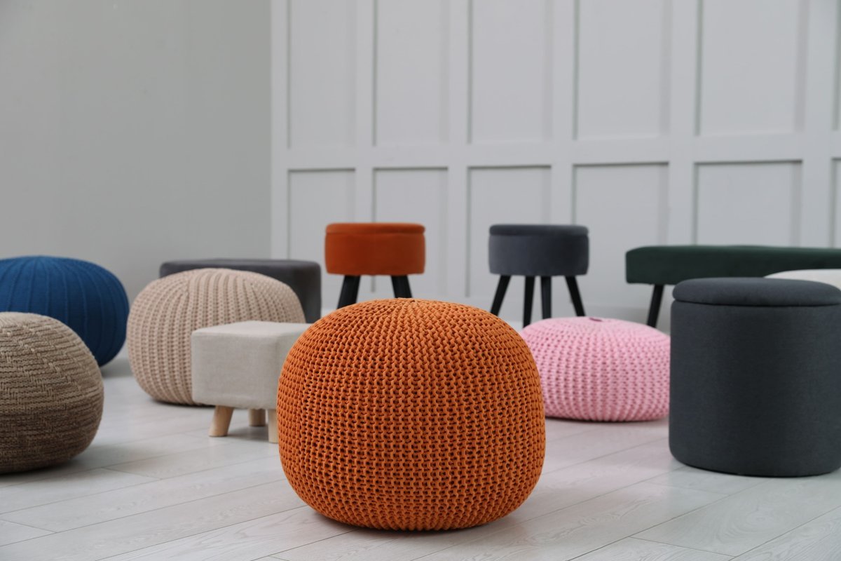 Various poufs, ottomans, and hassocks in different shapes, textures, and colors arranged in a modern room with light wooden flooring and white paneled walls.