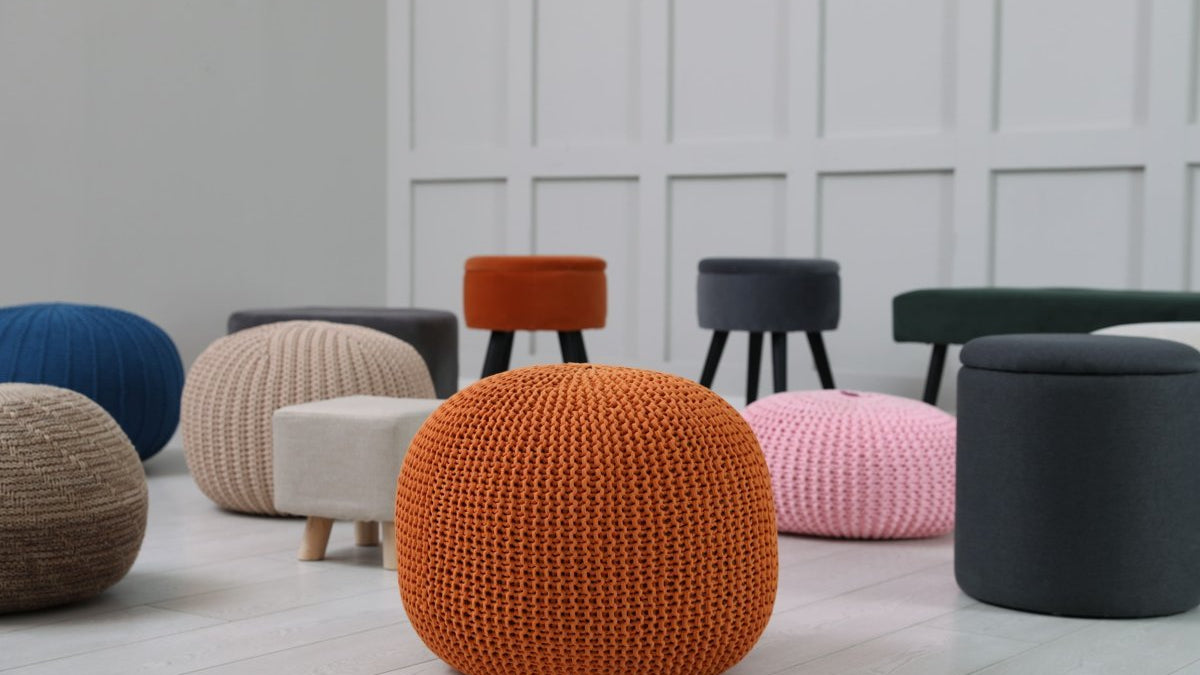 Various poufs, ottomans, and hassocks in different shapes, textures, and colors arranged in a modern room with light wooden flooring and white paneled walls.