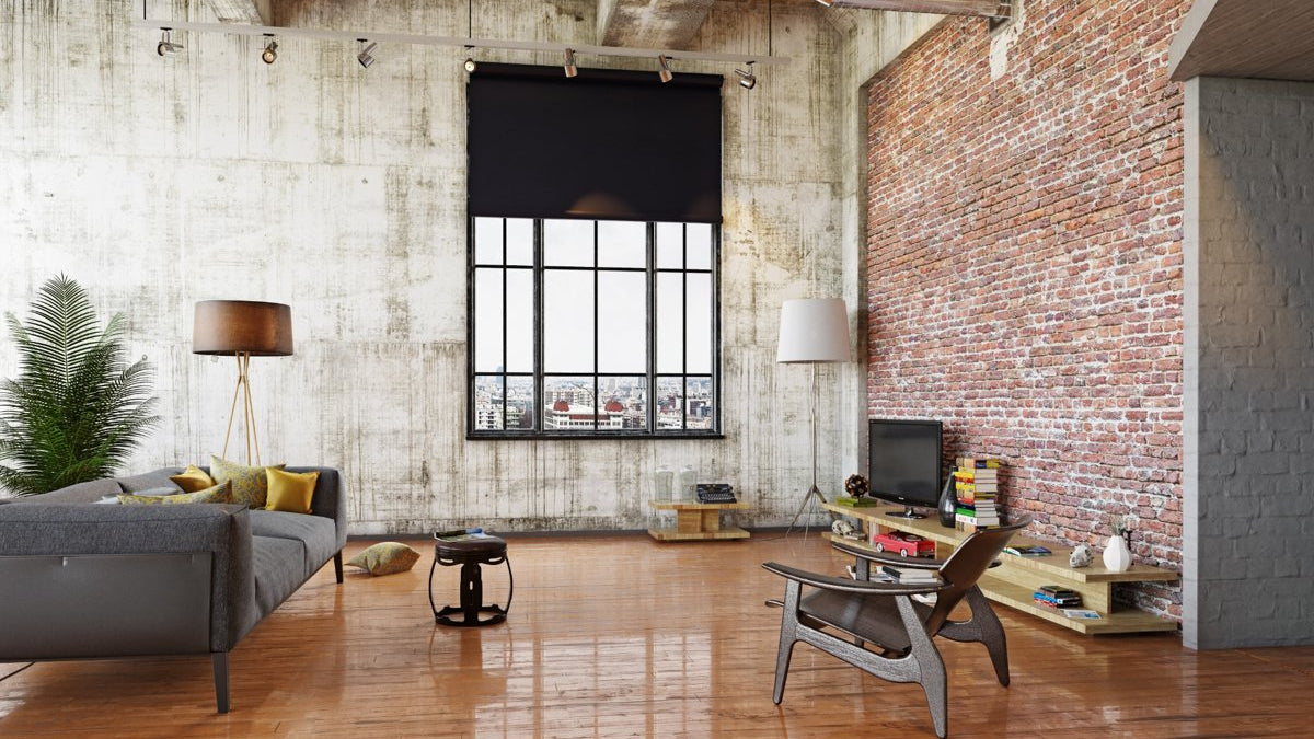 Industrial-style living room featuring exposed brick walls, a large window with a black roller blind, gray sofa with yellow cushions, wooden furniture, and polished wood flooring.