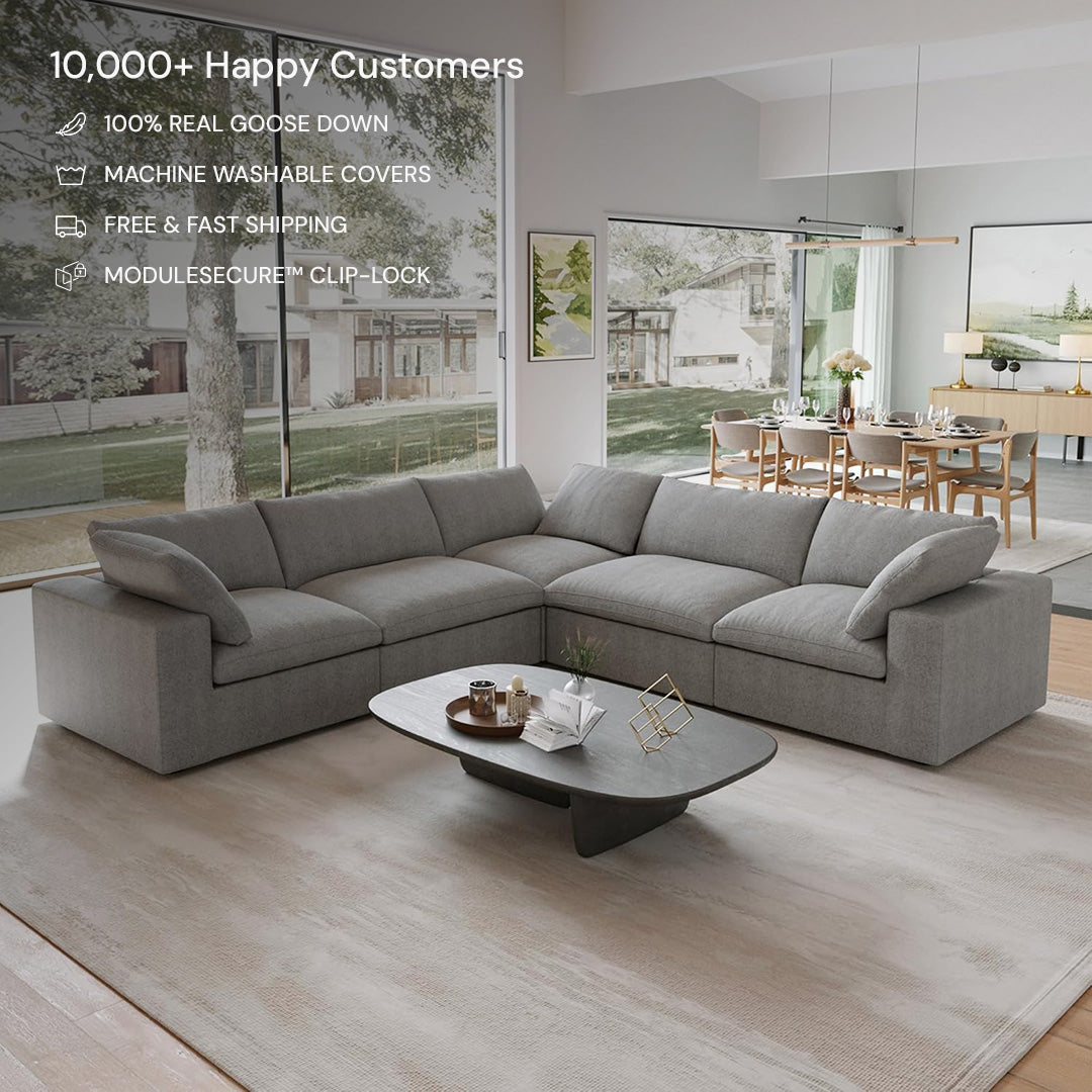 The bright living room showcases the CloudForm™ 5-Seater Modular L-Sofa in gray, featuring removable fabric covers, paired with a low coffee table on a large rug. Large windows and minimalistic decor complete the modern setting.